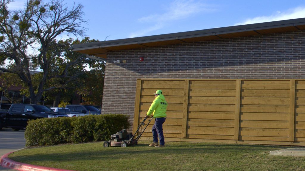 Landscape crew member mowing grass along commercial building with push mower