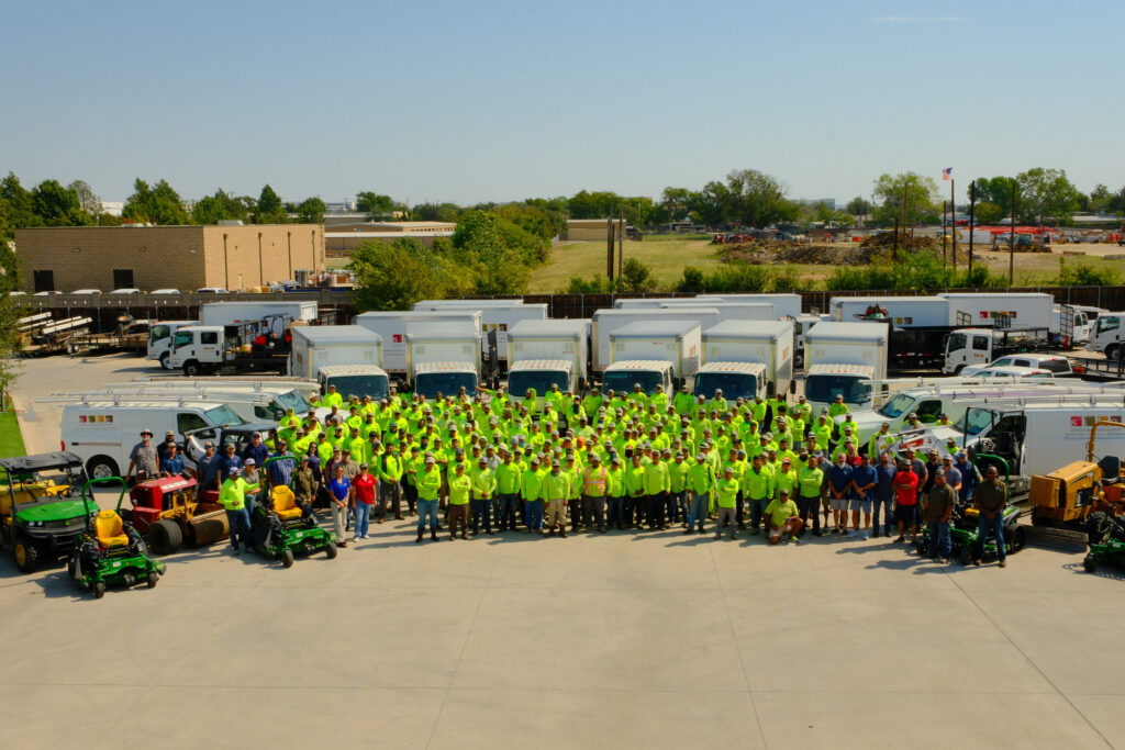 Aerial photo of the entire Site Landscape Development team in green shirts standing with their truck and mower fleet at the company headquarters in Lewisville, Texas.